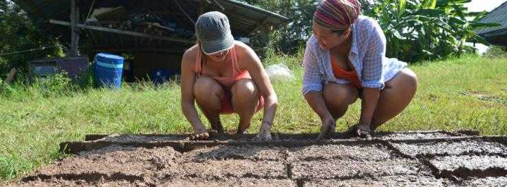 Two women prepare the materials for a new building on a building course. They are outside in the sun, and smiling.