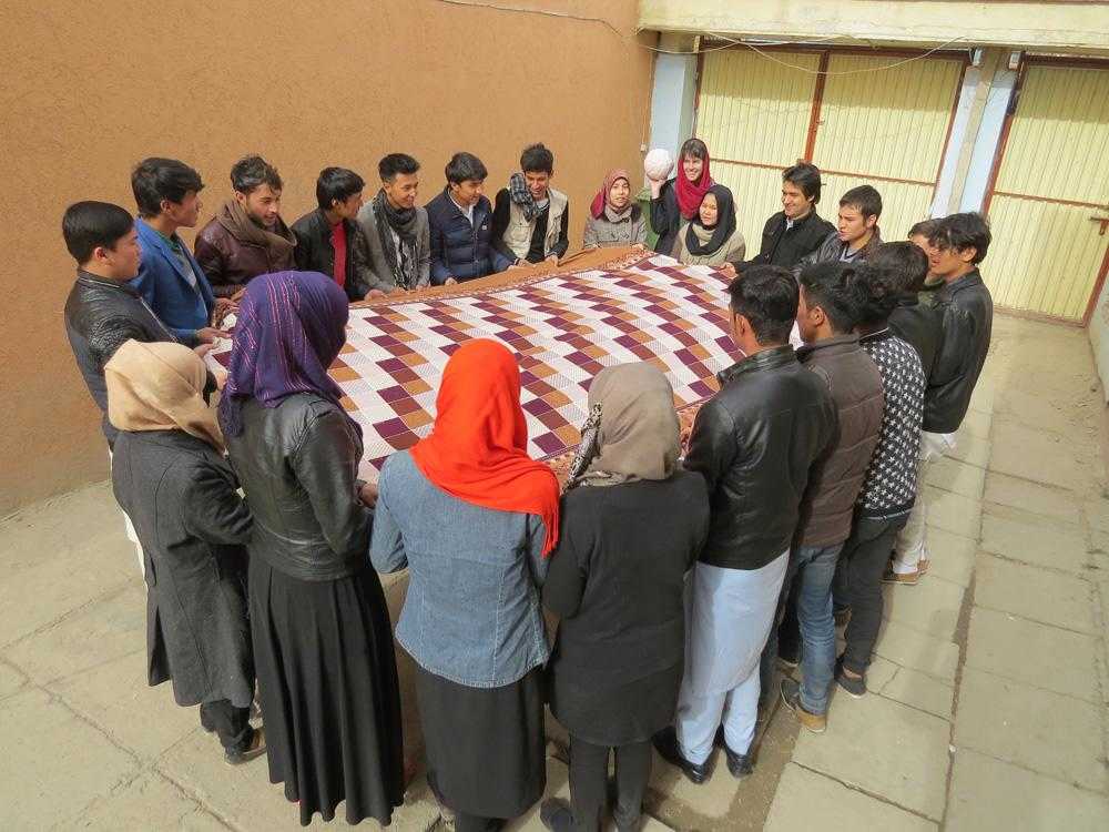 A dozen young people are gathered in a circle outside, holding a large multicoloured blanket between them. A woman prepares to throw a ball onto the blanket.
